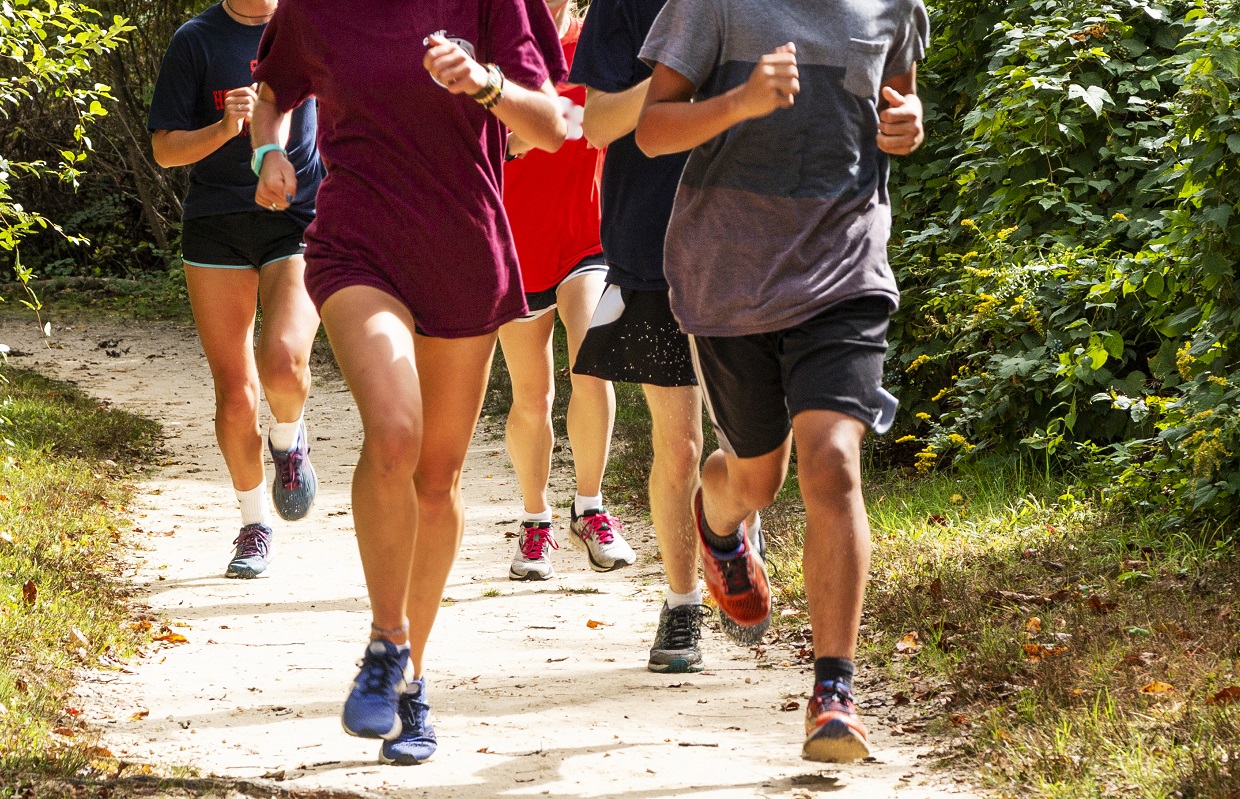 A group of high school runners are training together on a dirt path on a sunny afternoon, running toward the camera.